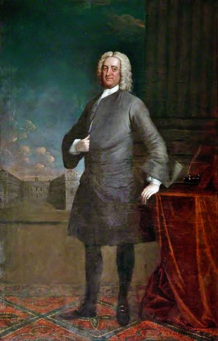 Portrait of Bryan Blundell, standing beside a draped table with a view of the Blue Coat School behind him